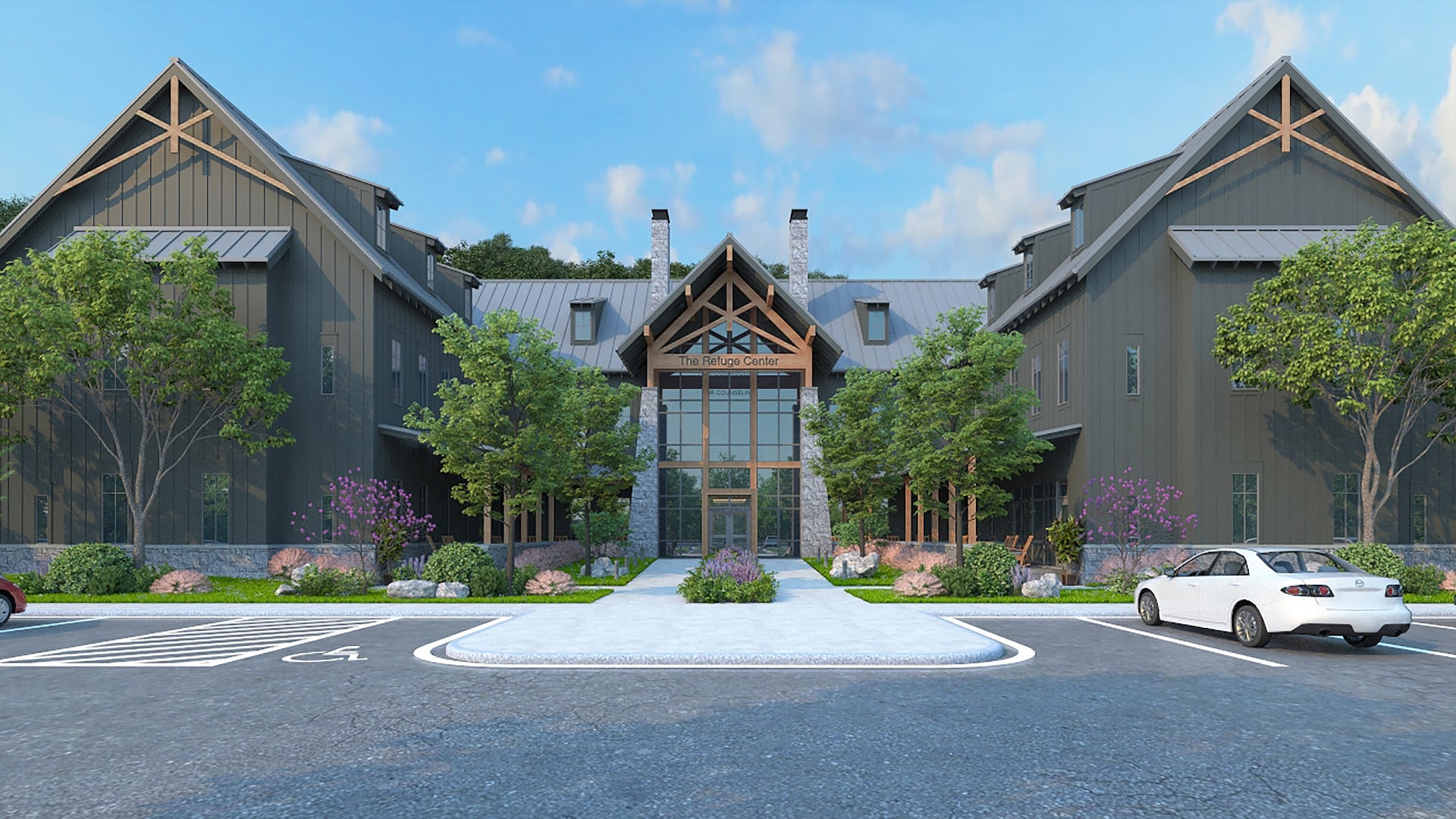 The image shows a large, modern building with a rustic design, featuring a central entrance with a high, peaked roof supported by wooden beams. The building is flanked by two symmetrical wings, each with a similar peaked roof. The exterior is clad in dark-colored siding, and there are several windows on each wing. In front of the building, there is a landscaped area with green grass, bushes, and small trees. A paved pathway leads to the entrance.