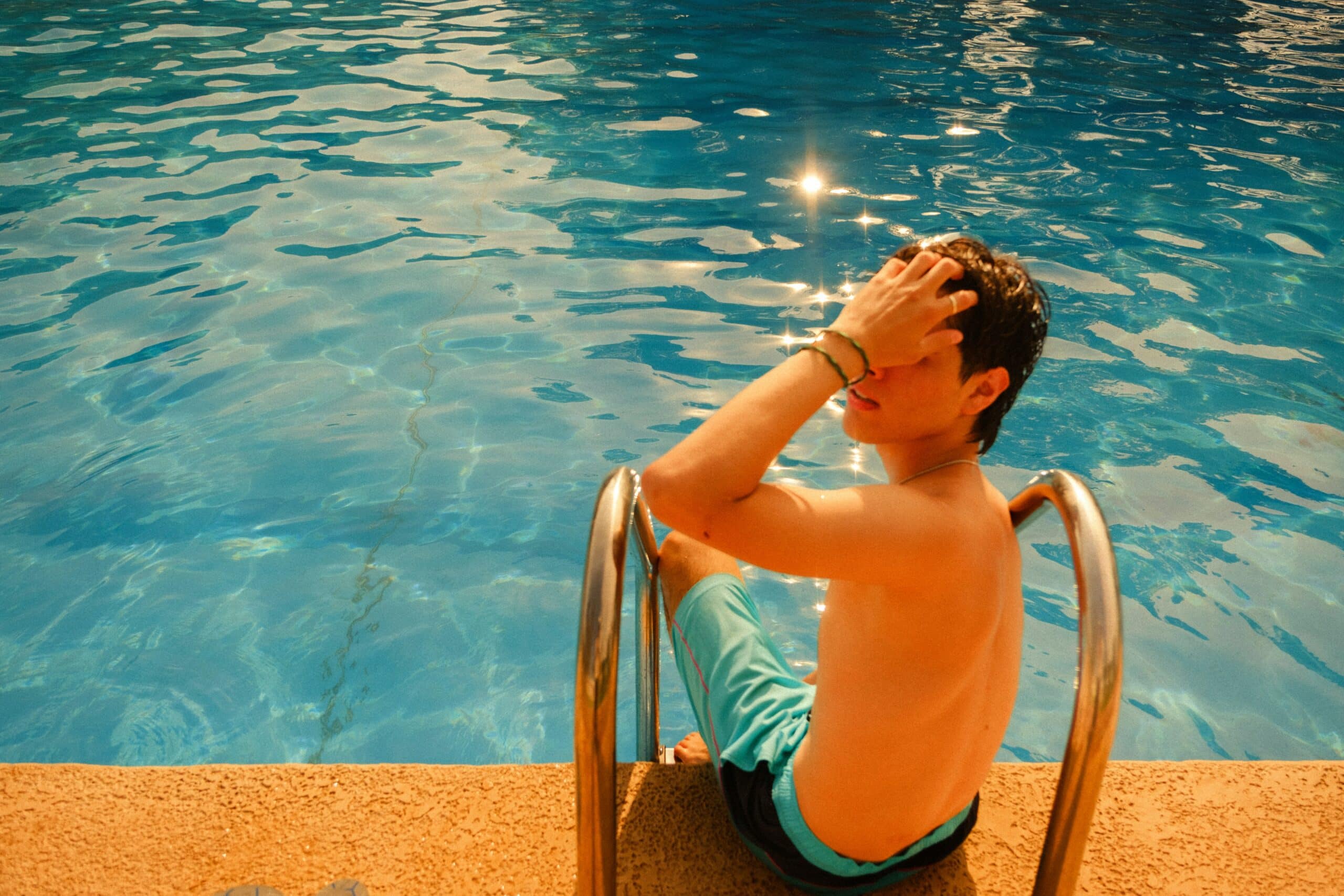 A boy sits by the pool, shielding his eyes